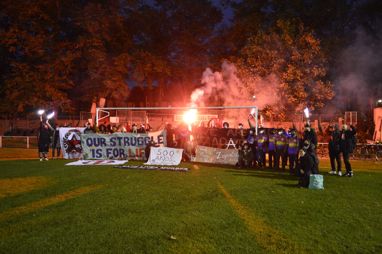 Group of people in a park holding flares and banners. Banners include messages such as "Our Struggle is For Life", "500 ANOS" and "We are all Zapatistas".